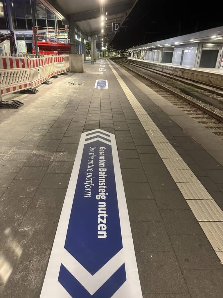 Frisch verklebte Bodenfolie „Gehen – Walk“ auf einem Bahnsteig, daneben eine Folienrolle für die nächste Markierung