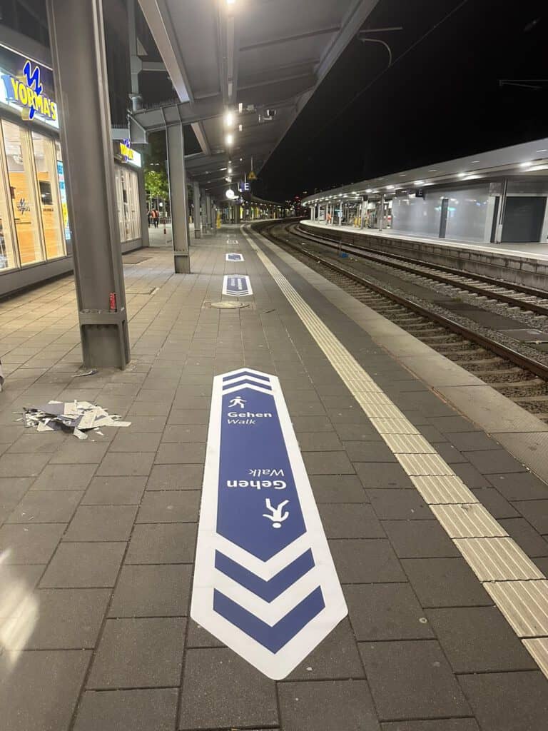 Bodenmarkierung „Gesamten Bahnsteig nutzen – Use the entire platform“ neben der Baustellenabsperrung an einem Bahnsteig im Münchner Hauptbahnhof