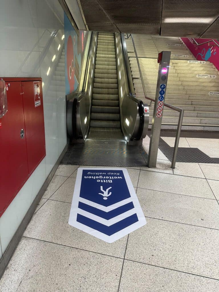 Bodenaufkleber „Bitte weitergehen“ am Fuß einer Rolltreppe im Münchner Hauptbahnhof