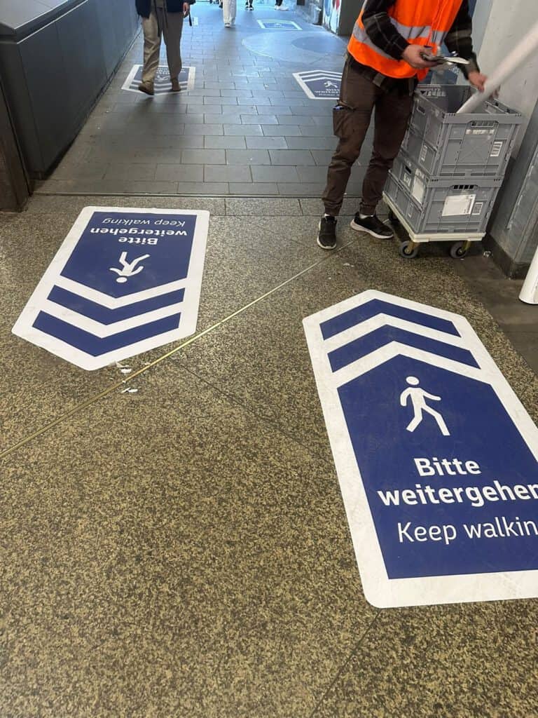 Monteur mit Materialkiste neben frisch verklebten Bodenaufklebern „Bitte weitergehen“ im Münchner Hauptbahnhof