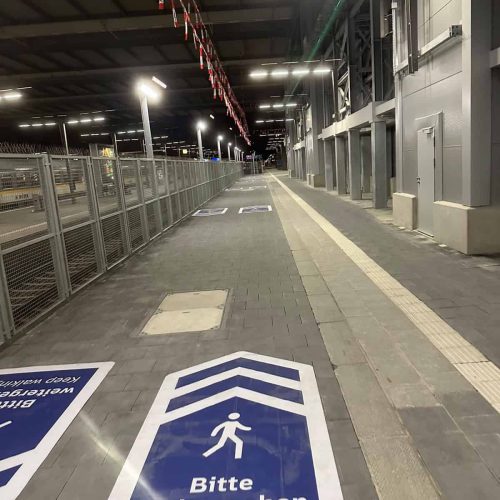 Bodenmarkierung „Bitte weitergehen – Keep walking“ auf einem Bahnsteig entlang eines Bauzauns im Münchner Hauptbahnhof
